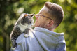 Smiling man carrying his curious cat on shoulder. Pet owner with old tabby cat in garden at sunny day..