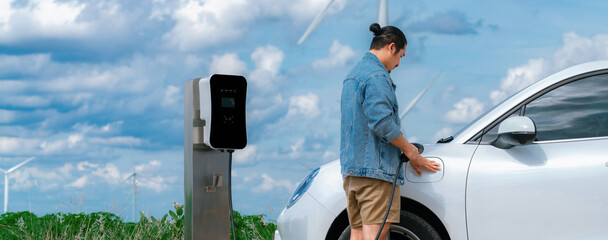 Progressive man with his electric car, EV car recharging energy from charging station on green field with wind turbine as concept of future sustainable energy. Electric vehicle with energy generator.