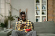 © JustLife - Handsome African man using smartphone while sitting on a sofa in cozy living room. Young man typing a message.