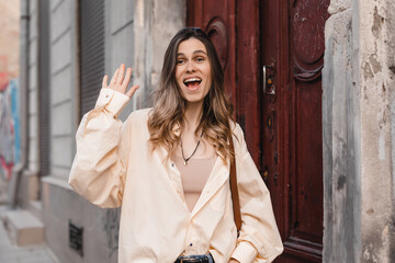 Wall Mural - Smiling young woman waving with a friendly cheerful smile to her new neighbours. Girl leaves the house standing near old door and waving her hand. Girl wear shirt and brown bag, meeting friends, hi.