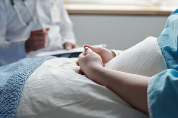 Close-up of hands of young woman with oncological disease sitting under blanket in bed and consulting with her doctor after medical treatment