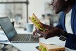 © pressmaster - Focus on appetizing vegetarian sandwich in hand of young hungry African American manager in formalwear using laptop at lunch break