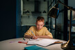 © dikushin - Portrait of concentrated pupil boy studying at home doing homework sitting at table under light of lamp at evening. Front view of schoolboy writing in exercise book at night in dark with lighting lamp