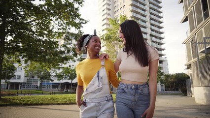 Wall Mural - Multiracial cute lesbian couple in love walk embracing laughing on city street. Two cheerful young friends stroll hugging on summer day in park. Gay women and relationships in youth generation z.