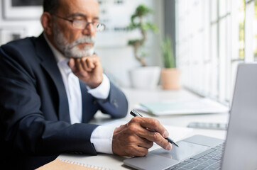 Serious confident pensive european senior businessman in suit looks at laptop with pen at table in office