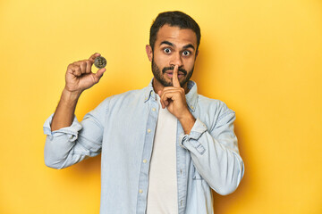 Wall Mural - Young Latino man holding a Bitcoin coin, yellow studio background, keeping a secret or asking for silence.