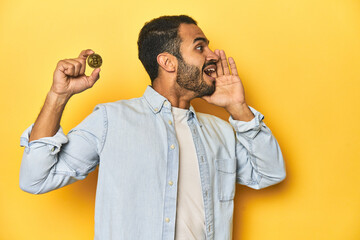 Wall Mural - Young Latino man holding a Bitcoin coin, yellow studio background, shouting and holding palm near opened mouth.