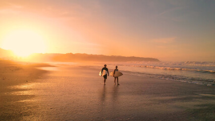 AERIAL: Young couple walking on beautiful sandy beach after sunset surf session