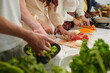 © DragonImages - Row of young intercultural friends with sharp knives chopping fresh vegetables for salad while standing by table and cooking dinner