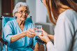 © VisualProduction - Nurse giving glass of water to elderly woman in wheelchair. Nursing home employee helping elderly people. Generative AI