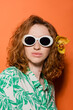 © LIGHTFIELD STUDIOS - Portrait of stylish redhead woman with orchid flower in hair posing in sunglasses and blouse with floral pattern on orange background, summer casual and fashion concept, Youth Culture