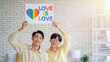 © Pitchayaarch - Happy gay Asian couple holding rainbow heart sign with 'Love Is Love' message indoors.