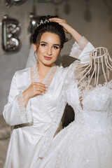 Wall Mural - A brunette bride in a satin robe stands near the dress. Beautiful hair and makeup. Wedding portrait. Sincere smile. A beautiful bride in a robe poses next to her dress