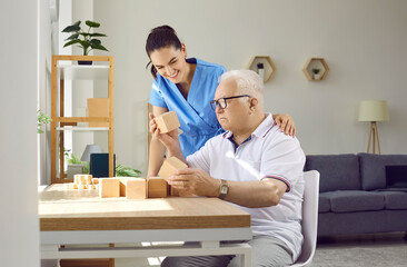 Wall Mural - Friendly nurse helping demented old man in retirement home. Senior man with cognitive problems sitting at desk and building wood block house. Dementia care, people with Alzheimer's disease concept