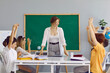© Studio Romantic - Children rising hands to answer teacher question sitting at desk in elementary school. Young woman tteacher is standing near desk looking at children and smiling. Education process in primary school.