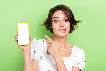 Photo of pretty unsure woman dressed white top biting lip pointing finger modern gadget empty space isolated green color background