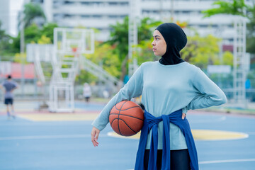 Wall Mural - Young asian muslim girl teen wearing hijab going to play basketball on the outdoor court in the morning with determination, Muslim sport concept.