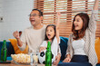 © Kiattisak - Asian Family with daughter watching sport football cheering excited eating popcorn on sofa in living room at home. enjoy relaxing happiness.