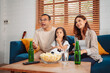 © Kiattisak - Asian Family with daughter watching sport football cheering excited eating popcorn on sofa in living room at home. enjoy relaxing happiness.