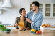 © Kateryna - Multiracial happy couple in casual wear, young spouses, stand at home in the kitchen, prepare salad together, woman cuts vegetables, man hugs her, they look at each other with love, smile