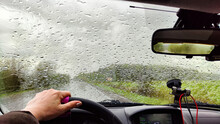 View From The Car Windshield From The Driver's Seat And Hand On The Steering Wheel Of Woman On During Rain, Drops On The Glass And Nature. Travel In Rainy Autumn, Spring Or Summer Weather