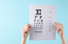 Hands Holding Eye Test Chart Paper Sheet On Blue Background. Vision Correction, Visit To Ophthalmologist