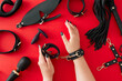 Experience passion with alluring BDSM toys. First person top view of female hands in black leather handcuffs and different sex toys for role play on red background