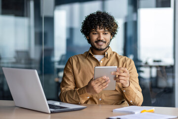 Young successful businessman at workplace is satisfied with results of achievement at work smiling and using tablet computer, hispanic programmer is testing new software in application