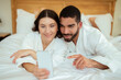 © Prostock-studio - Cheerful Couple Using Smartphone Lying In Bed In Hotel Indoors