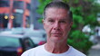 © Marco - Portrait of a serious middle-aged man standing in city street looking at camera with neutral expression. Close-up face of a male older person in urban environment