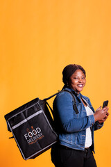 Wall Mural - Restaurant delivery employee holding smartphone looking at customer adreess on fast food app before start deliver takeout order. Pizzeria courier carrying takeaway thermal backpack in studio