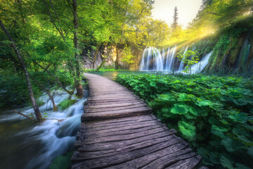 Waterfall and wooden path in green forest in Plitvice Lakes, Croatia at sunset in summer. Colorful landscape with trail in blooming park, trees, water lilies, river, sunbeams in spring. Trail in woods