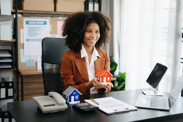 Young real estate agent worker working with laptop and tablet at table in office and small house beside it.