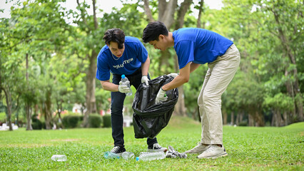 Two happy Asian men with garbage bag helping each other clean up the area in a public park.