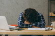 © ViDi Studio - Sad tired exhausted sleepless employee business Indian man he wearing casual blue checkered shirt sleeping taking nap crying put head on table sit work at office desk with laptop pc computer indoors.