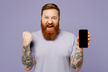 Young redhead bearded man wearing violet t-shirt casual clothes hold in hand use mobile cell phone with blank screen workspace area do winner gesture isolated on plain pastel light purple background.