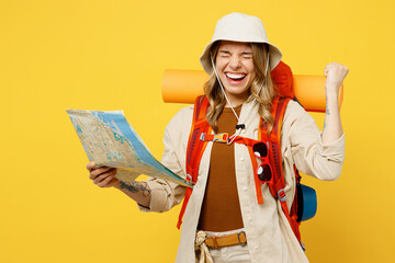 Young happy woman carry backpack with stuff mat hold map do winner gesture isolated on plain yellow background. Tourist leads active lifestyle walk on spare time. Hiking trek rest travel trip concept.