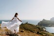 © svetograph - Happy woman in a white dress and hat stands on a rocky cliff above the sea, with the beautiful silhouette of hills in thick fog in the background.