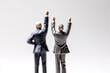 Miniature people figurines of two excited businessmen dressed in formal suits stand with their hands raised in celebrating gesture on isolated background, created with Generative AI