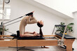 © Georgii - A young girl is doing Pilates on a reformer bed in a bright studio. A slender brunette in a beige bodysuit does exercises to strengthen the arms and shoulder girdle. Healthy lifestyle concept.