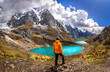 Man standing in front of a teal water lake with high Peruvian mountains in the Huayhuash mountain range, Peru