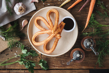 Fresh baked sweet churros with chocolate sauce on a plate on rustic wooden table, ingredients surrounding the plate, top view