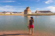 Rear view of woman walking at lakeshore looking at solitary rock formation Lone Rock in Wahweap Bay at Lake Powell, Glen Canyon Recreation Area, Page, Utah, USA. Lone rock campground in sunny summer