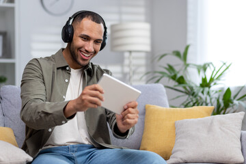 A young Hispanic man plays online games on a tablet at home. Sitting smiling on the couch wearing headphones