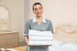 Young chambermaid holding stack of clean towels in hotel room