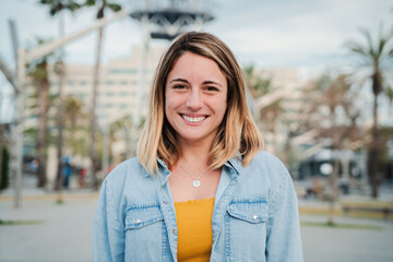 Individual portrait of happy blonde young adult woman standing outside with friendly and optimistic expression. Caucasian beauty female with perfect white teeth smiling and looking satisfed at camera