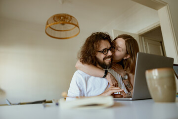 Wall Mural - Young smiling man working on laptop while girlfriend is kissing him at home. Distance work concept