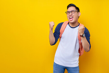 Wall Mural - Excited young Asian man student wearing t-shirt with backpack making winner gesture and celebrating success isolated on yellow background. Education in high school university college concept
