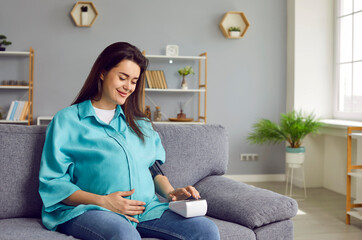 Wall Mural - Happy pregnant woman measures blood pressure. Beautiful smiling young lady sitting on sofa, touching big belly and using tonometer to check her normal good arterial pressure. Pregnancy, health concept