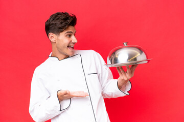 Wall Mural - Young caucasian chef man holding tray isolated on red background with surprise expression while looking side